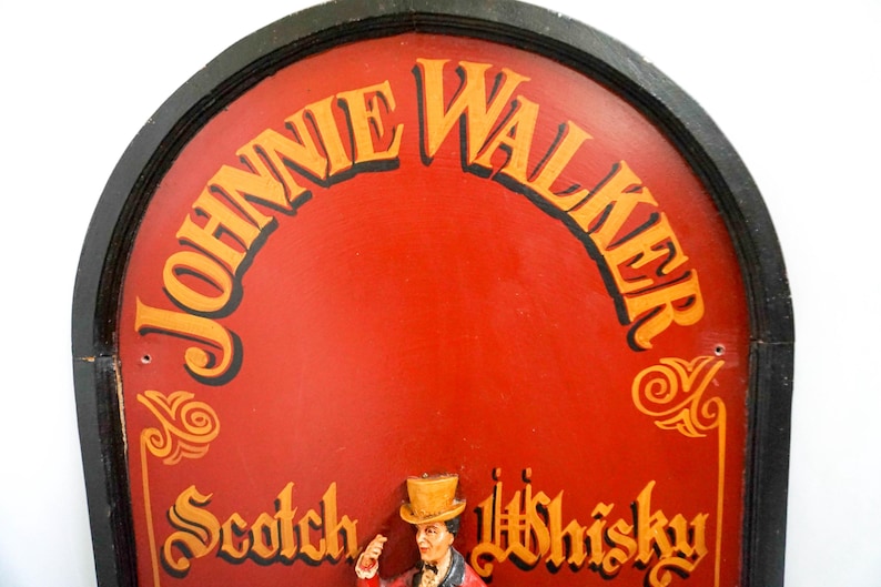 May include: A vintage wooden sign with a red background and black trim. The sign features the text "Johnnie Walker Scotch Whisky" in gold lettering. The sign is shaped like an arch and has a decorative design around the edges.