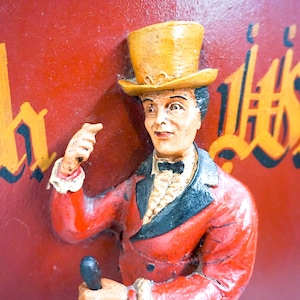 May include: A figurine of a person wearing a yellow top hat and a red jacket with a black collar. The figurine is holding a small object in its hand. The background is red with the words "Rich" and "Whisky" written in gold.