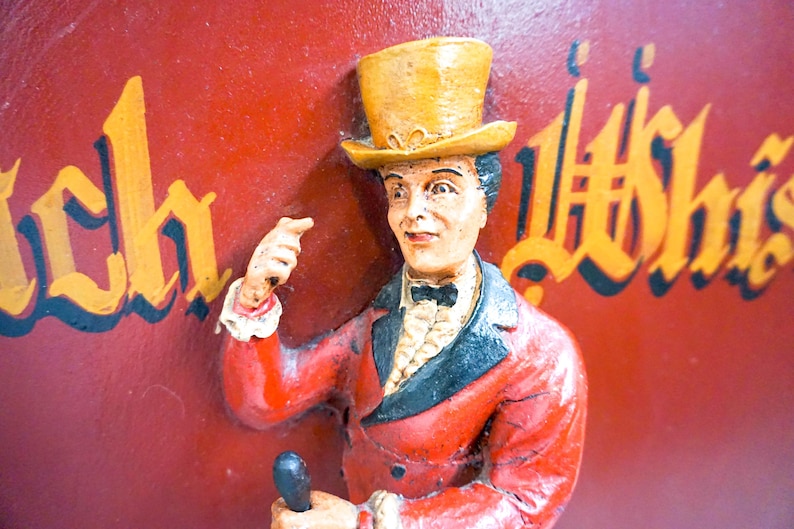May include: A figurine of a person wearing a yellow top hat and a red jacket with a black collar. The figurine is holding a small object in its hand. The background is red with the words "Rich" and "Whisky" written in gold.