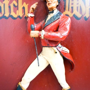 May include: A vintage sign featuring a man in a red jacket and white pants holding a stick. The sign has the words "Scotch Whisky" in gold lettering.