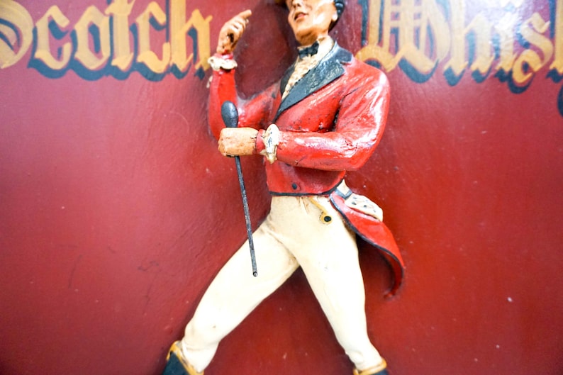 May include: A vintage sign featuring a man in a red jacket and white pants holding a stick. The sign has the words "Scotch Whisky" in gold lettering.