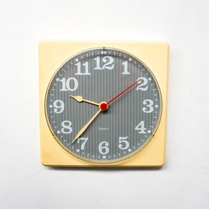 Puede incluir: Un reloj redondo de color crema con números negros y una esfera gris. El reloj tiene una aguja de segundos roja y las palabras "Quartz" impresas en la esfera.