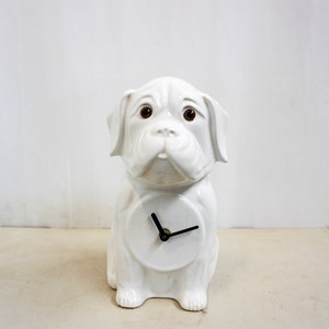 Op de afbeelding: Een witte keramische klok in de vorm van een hond, met bruine ogen en zwarte wijzers. De klok heeft een ronde wijzerplaat en zit rechtop. De hond heeft een vriendelijke uitdrukking en is een decoratief item.