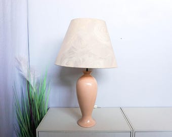 May include: A peach-coloured ceramic table lamp with a cream-coloured lampshade. The lamp has a vase-like base and a tapered shade. The lamp is on a grey surface, with green and white plants to the left.