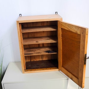 Op de afbeelding: Een houten wandkast met een enkele deur en vier planken. De deur staat open en de kast is leeg.