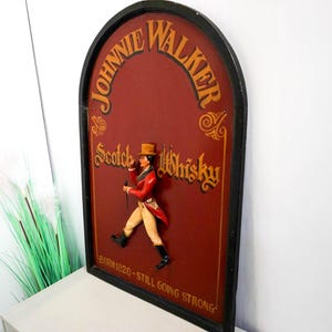 May include: A vintage wooden sign with a red background and gold lettering. The sign features a 3D figure of a man in a red coat and black hat, holding a walking stick. The sign reads "Johnnie Walker Scotch Whisky - Born 1820 - Still Going Strong."