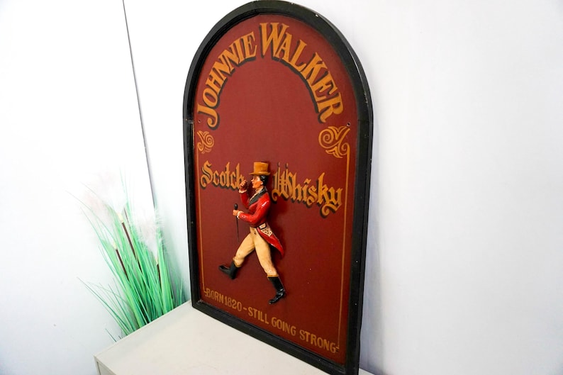 May include: A vintage wooden sign with a red background and gold lettering. The sign features a 3D figure of a man in a red coat and black hat, holding a walking stick. The sign reads "Johnnie Walker Scotch Whisky - Born 1820 - Still Going Strong."