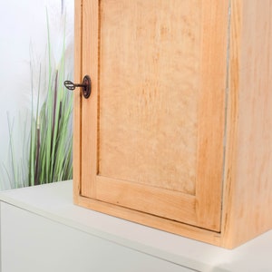 Op de afbeelding: Een licht houten kast met een enkele deur en een sleutelgat. De kast heeft een eenvoudig ontwerp en is gemonteerd op een witte muur.