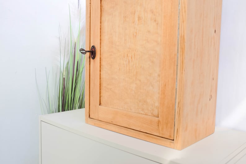 Op de afbeelding: Een licht houten kast met een enkele deur en een sleutelgat. De kast heeft een eenvoudig ontwerp en is gemonteerd op een witte muur.