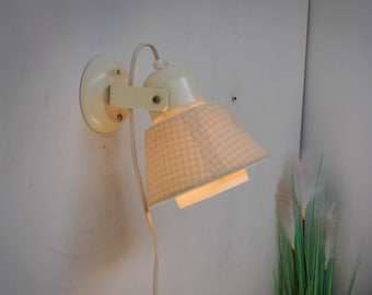 Puede incluir: Una lámpara de pared blanca con una pantalla de vichy blanca y rosa. La lámpara está enchufada y tiene un cable.