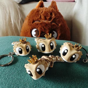 Pode incluir: Quatro figuras de lagarto articuladas em tons de bege e branco com grandes olhos pretos e brancos, cada uma usando uma pequena coroa dourada. Uma figura está presa a um porta-chaves de metal prateado e outra a um cordão preto com fecho.