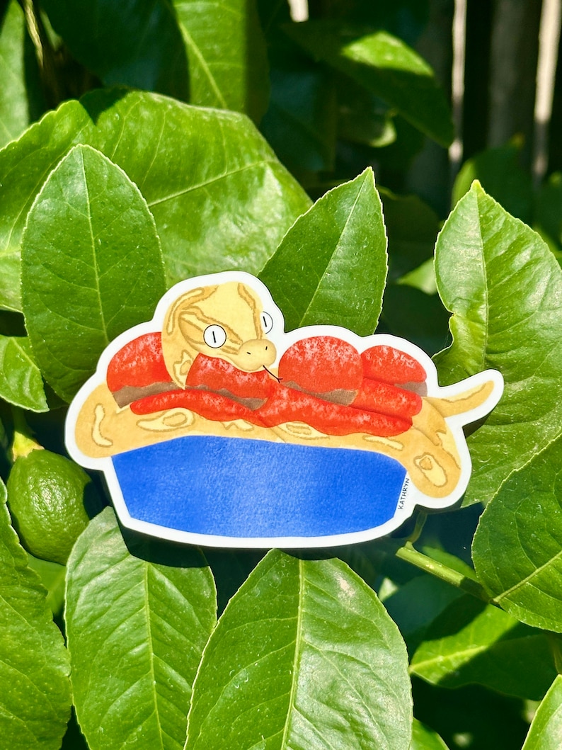 K&ouml;nnte beinhalten: Ein Aufkleber mit einer Cartoon-Schlange in einer blauen Sch&uuml;ssel mit roten und braunen Bel&auml;gen. Die Schlange ist gelb mit braunen Flecken und hat ein wei&szlig;es Auge. Der Aufkleber befindet sich auf einem gr&uuml;nen, bl&auml;ttrigen Hintergrund.