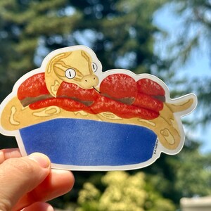 K&ouml;nnte beinhalten: Ein Aufkleber mit einer Cartoon-Schlange mit einem gelben und braunen K&ouml;rper, die sich in einer roten Sch&uuml;ssel zusammenrollt. Die Schlange hat einen hellbraunen Kopf mit einem wei&szlig;en Streifen und zwei gro&szlig;en schwarzen Augen. Die Sch&uuml;ssel ist blau mit einem wei&szlig;en Rand. Der Aufkleber ist auf wei&szlig;em Hintergrund.