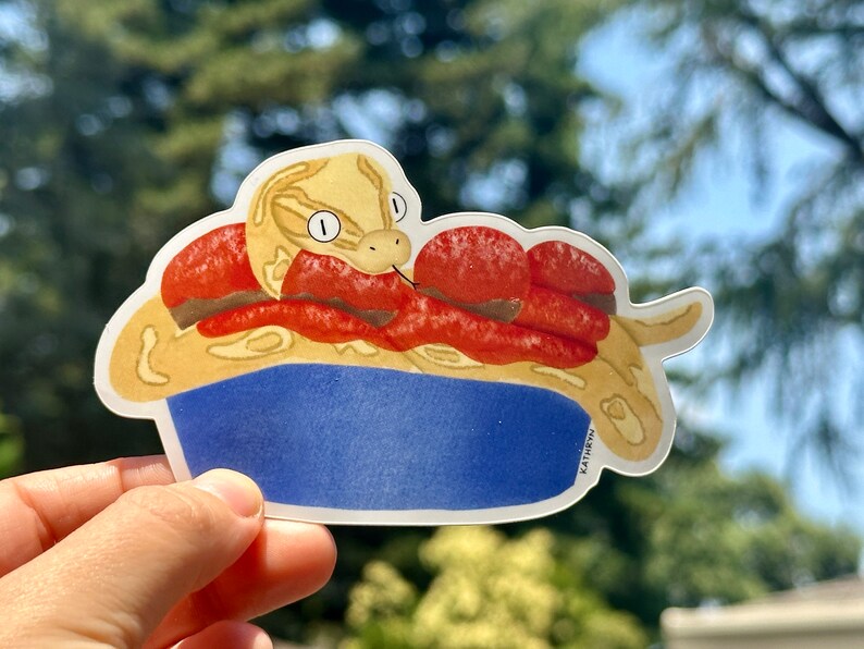 K&ouml;nnte beinhalten: Ein Aufkleber mit einer Cartoon-Schlange mit einem gelben und braunen K&ouml;rper, die sich in einer roten Sch&uuml;ssel zusammenrollt. Die Schlange hat einen hellbraunen Kopf mit einem wei&szlig;en Streifen und zwei gro&szlig;en schwarzen Augen. Die Sch&uuml;ssel ist blau mit einem wei&szlig;en Rand. Der Aufkleber ist auf wei&szlig;em Hintergrund.