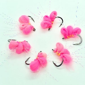 May include: Six pink fly fishing lures with white and iridescent fibers. The lures have a hook and are tied with pink and yellow yarn.