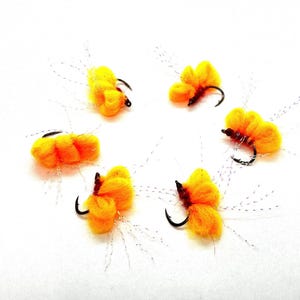 May include: Six bright yellow and orange fishing flies with black hooks and silver thread accents are arranged on a white background. The flies have a fluffy, textured appearance, suggesting they are made of yarn or similar material.
