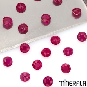 Puede incluir: Una colección de 16 piedras preciosas redondas de color rojo, probablemente rubíes, esparcidas sobre una superficie blanca. Algunas de las piedras preciosas están en una bandeja plateada.