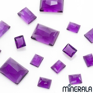 May include: A collection of twelve amethyst gemstones, cut into rectangular shapes, displayed on a white surface. The gemstones are various sizes and have a deep purple hue.
