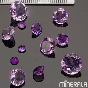 Puede incluir: Una colección de gemas de amatista de varios tamaños y formas, que van desde la redonda hasta la ovalada, expuestas sobre una superficie oscura. Las gemas son de un color morado vibrante.