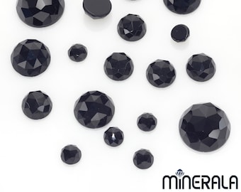 May include: A collection of black, faceted gemstones of varying sizes, arranged on a white surface. The gemstones have a polished, reflective surface, and the facets create a play of light and shadow. The image includes a logo in the bottom right corner.