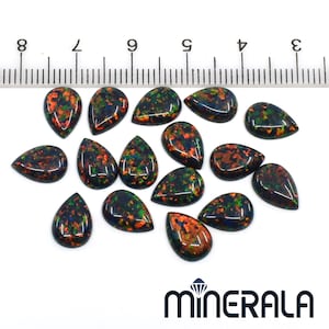 May include: A collection of teardrop-shaped black opals with vibrant red, orange, and green flashes. The opals are arranged on a white surface next to a ruler, with the "MINERALA" logo visible.