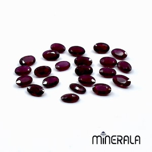 Puede incluir: Una colección de 20 gemas ovaladas de color rojo oscuro. Las gemas están esparcidas sobre una superficie blanca.