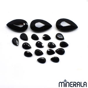 Puede incluir: Una colecci&oacute;n de gemas negras pulidas en forma de l&aacute;grima de varios tama&ntilde;os. Las gemas est&aacute;n dispuestas sobre una superficie blanca, con tres piedras grandes en la parte superior y piedras m&aacute;s peque&ntilde;as debajo. La imagen est&aacute; bien iluminada, resaltando las superficies reflectantes.