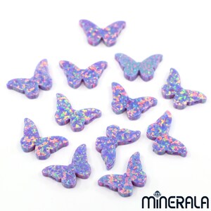 Puede incluir: Una colecci&oacute;n de 12 abalorios de mariposa de &oacute;palo p&uacute;rpura iridiscente. Los abalorios est&aacute;n esparcidos sobre un fondo blanco.