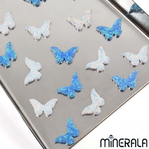 Peut inclure: Assortiment de breloques papillon en opale iridescente bleue et blanche. Les breloques sont éparpillées sur une surface argentée.