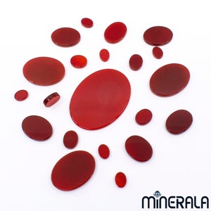Peut inclure: Plusieurs pierres précieuses rouges polies, de forme ovale et ronde, de différentes tailles, sont disposées sur un fond blanc. Les pierres ont une finition brillante et sont disposées de manière éparse. Le logo "MINERALA" est visible en bas à droite.