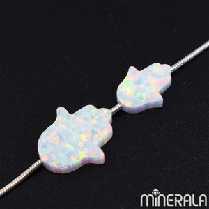 Puede incluir: Dos amuletos de mano de Hamsa de ópalo blanco en una cadena de plata. Los amuletos son iridiscentes y tienen un aspecto moteado.