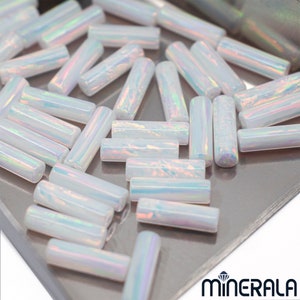 May include: A collection of iridescent white opal beads in a cylindrical shape. The beads are arranged on a silver surface.