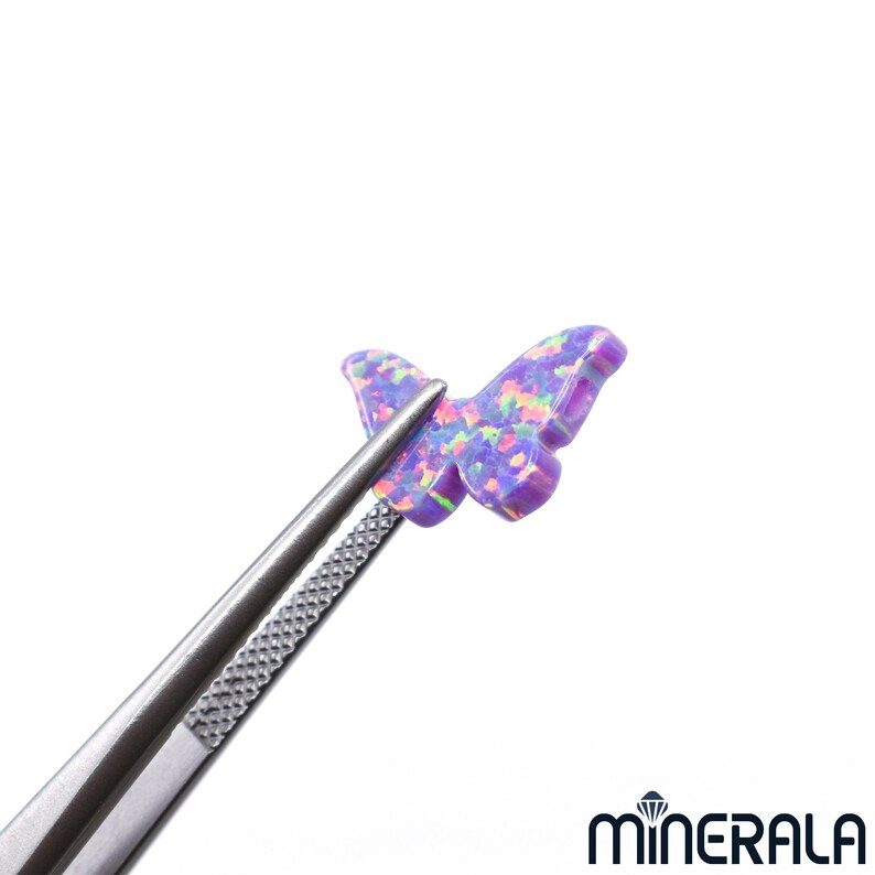 Puede incluir: Un cabuj&oacute;n de &oacute;palo de forma de mariposa de color p&uacute;rpura con colores arco&iacute;ris iridiscentes. El &oacute;palo est&aacute; sujeto por unas pinzas.