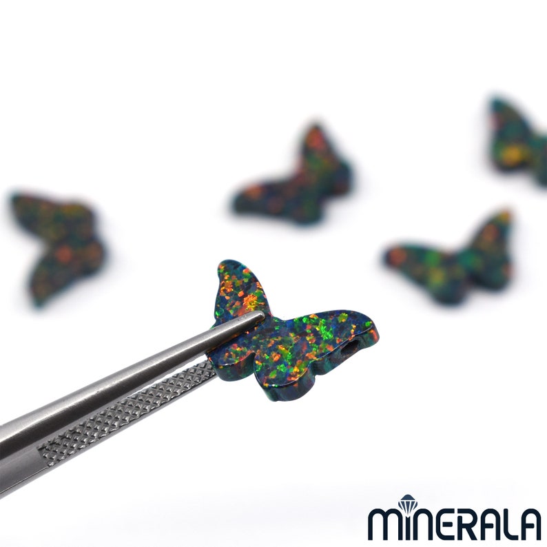 Puede incluir: Primer plano de un dije de mariposa de &oacute;palo negro con colores iridiscentes verdes, naranjas y rojos. El dije se sostiene con unas pinzas sobre un fondo blanco.