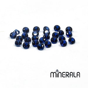 Puede incluir: Una colección de 20 piedras preciosas redondas, facetadas, de color azul oscuro. Las piedras preciosas están dispuestas en un patrón suelto y disperso sobre una superficie blanca.