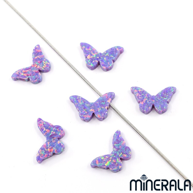 Puede incluir: Seis abalorios de mariposa de &oacute;palo p&uacute;rpura con brillo iridiscente. Los abalorios est&aacute;n dispuestos sobre una superficie blanca con una cadena plateada.