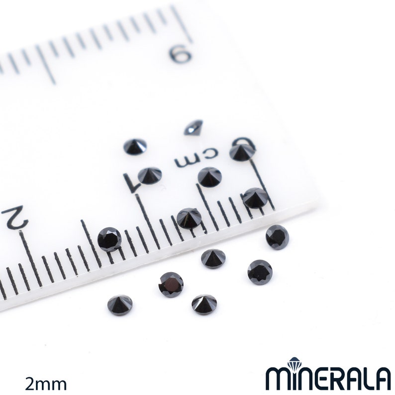 May include: A close-up of black diamond gemstones, measuring 2 mm in diameter, scattered on a white surface next to a ruler. The ruler is marked in centimetres and inches.
