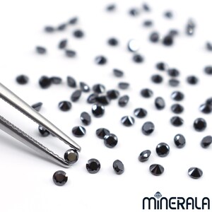 May include: A close-up of a pair of tweezers holding a black diamond gemstone. Several other black diamonds are scattered on a white surface.