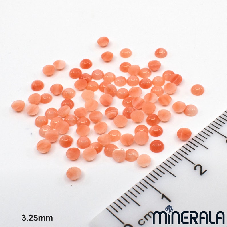 Pu&ograve; includere: Una collezione di circa 50 pietre preziose rotonde in cabochon rosa e bianco, che misurano 3,25 mm di diametro. Le pietre preziose sono sparse su una superficie bianca accanto a un righello.