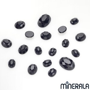Peut inclure: Une collection de pierres pr&eacute;cieuses ovales noires, certaines avec des bords facet&eacute;s, &eacute;parpill&eacute;es sur une surface blanche.