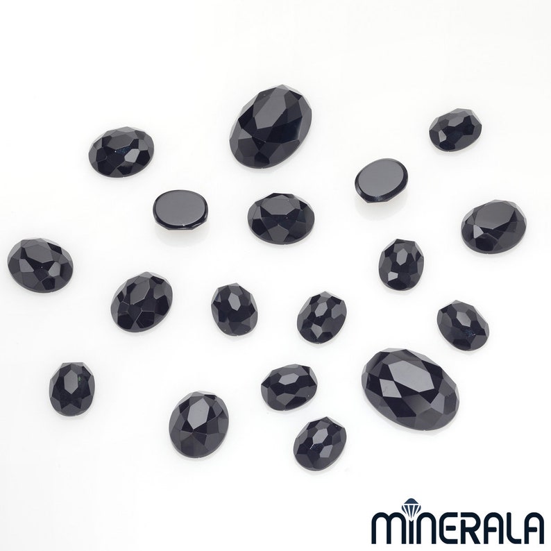 Peut inclure: Une collection de pierres pr&eacute;cieuses ovales noires, certaines avec des bords facet&eacute;s, &eacute;parpill&eacute;es sur une surface blanche.
