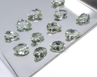 May include: A collection of pear-shaped, pale green gemstones displayed on a silver tray. The faceted stones are arranged in a scattered pattern, showcasing their clarity and cut. The logo "MINERALA" is visible in the bottom right corner.