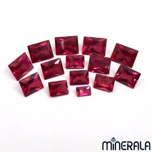 May include: A collection of radiant, square and rectangular-cut, deep red gemstones. The gems are arranged on a white surface, showcasing their facets and vibrant color. The image highlights the variety of sizes and shapes.