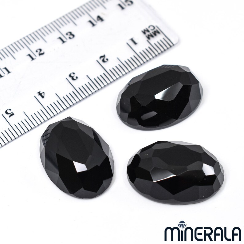 Peut inclure: Trois pierres pr&eacute;cieuses noires de forme ovale avec des bords facet&eacute;s. Les pierres pr&eacute;cieuses mesurent environ 2,5 cm de long et sont pr&eacute;sent&eacute;es sur une surface blanche &agrave; c&ocirc;t&eacute; d'une r&egrave;gle.