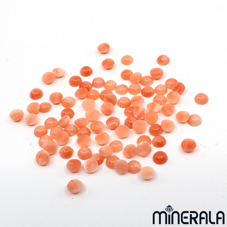 Pu&ograve; includere: Una collezione di circa 50 cabochon rotondi di colore pesca. I cabochon sono lisci e lucidi, con un leggero bagliore. Sono sparsi su una superficie bianca.