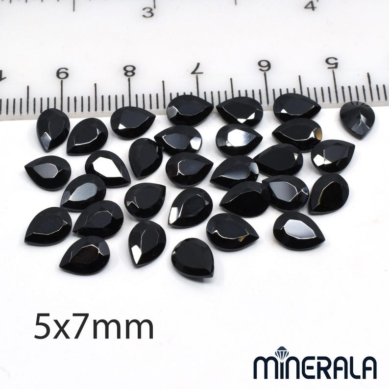 Puede incluir: Colecci&oacute;n de gemas negras brillantes en forma de l&aacute;grima. Las gemas est&aacute;n dispersas sobre una superficie blanca, con una regla en el fondo. El texto "5x7mm" es visible. Las gemas miden aproximadamente 5 x 7 mm.