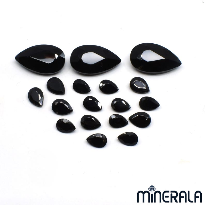 Puede incluir: Una colecci&oacute;n de gemas negras pulidas en forma de l&aacute;grima de varios tama&ntilde;os. Las gemas est&aacute;n dispuestas sobre una superficie blanca, con tres piedras grandes en la parte superior y piedras m&aacute;s peque&ntilde;as debajo. La imagen est&aacute; bien iluminada, resaltando las superficies reflectantes.