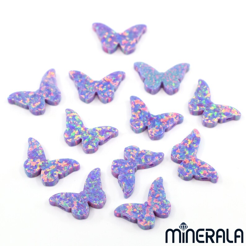 Puede incluir: Una colecci&oacute;n de 12 abalorios de mariposa de &oacute;palo p&uacute;rpura iridiscente. Los abalorios est&aacute;n esparcidos sobre un fondo blanco.