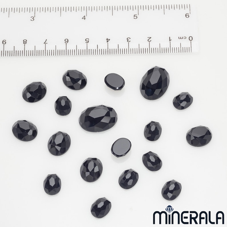 Peut inclure: Une collection de 17 pierres pr&eacute;cieuses ovales noires de tailles diff&eacute;rentes. Les pierres pr&eacute;cieuses sont facett&eacute;es et ont une finition brillante. Les pierres pr&eacute;cieuses sont dispos&eacute;es sur une surface blanche.