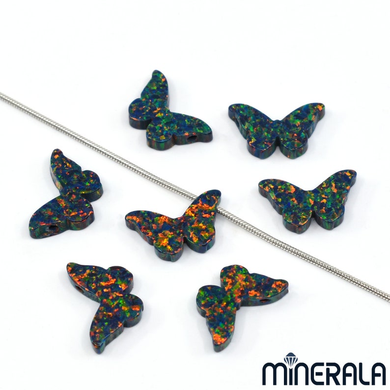 Puede incluir: Siete abalorios de mariposa de &oacute;palo iridiscente con una cadena plateada en el fondo. Las mariposas son de color azul oscuro con acentos iridiscentes verdes, naranjas y rojos.